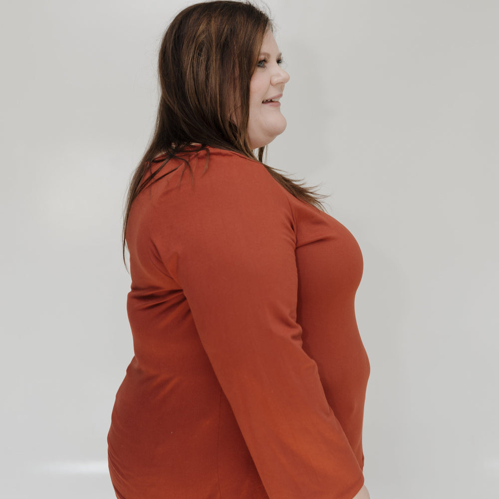 Woman wearing an orange long-sleeve top and blue jeans against a white wall.