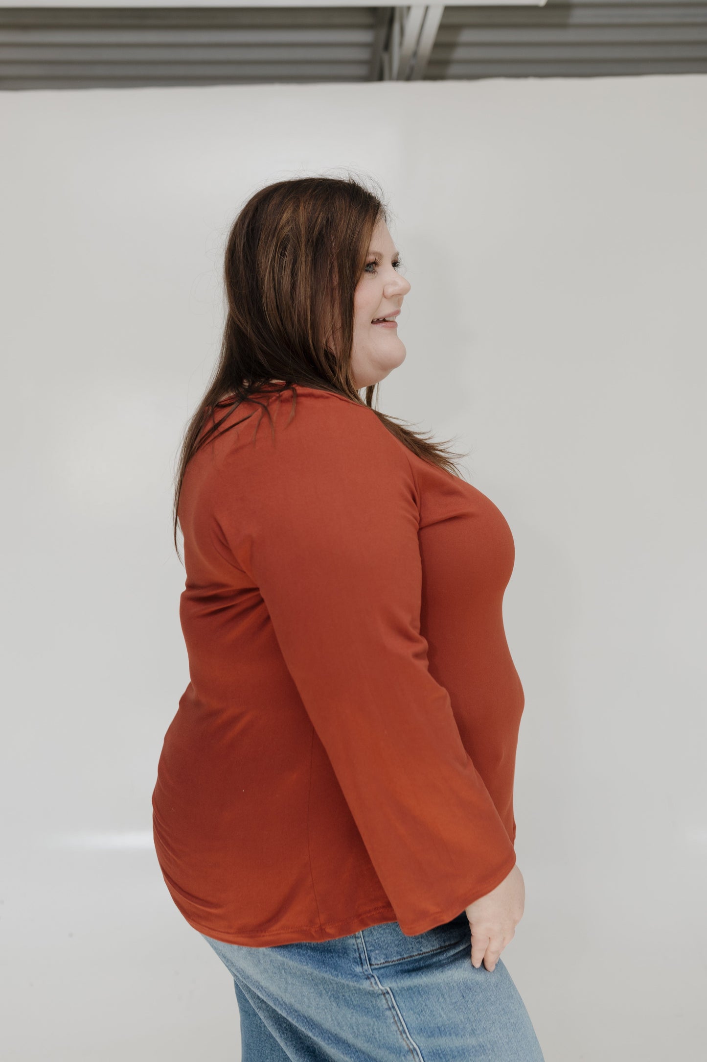 Woman wearing an orange long-sleeve top and blue jeans against a white wall.