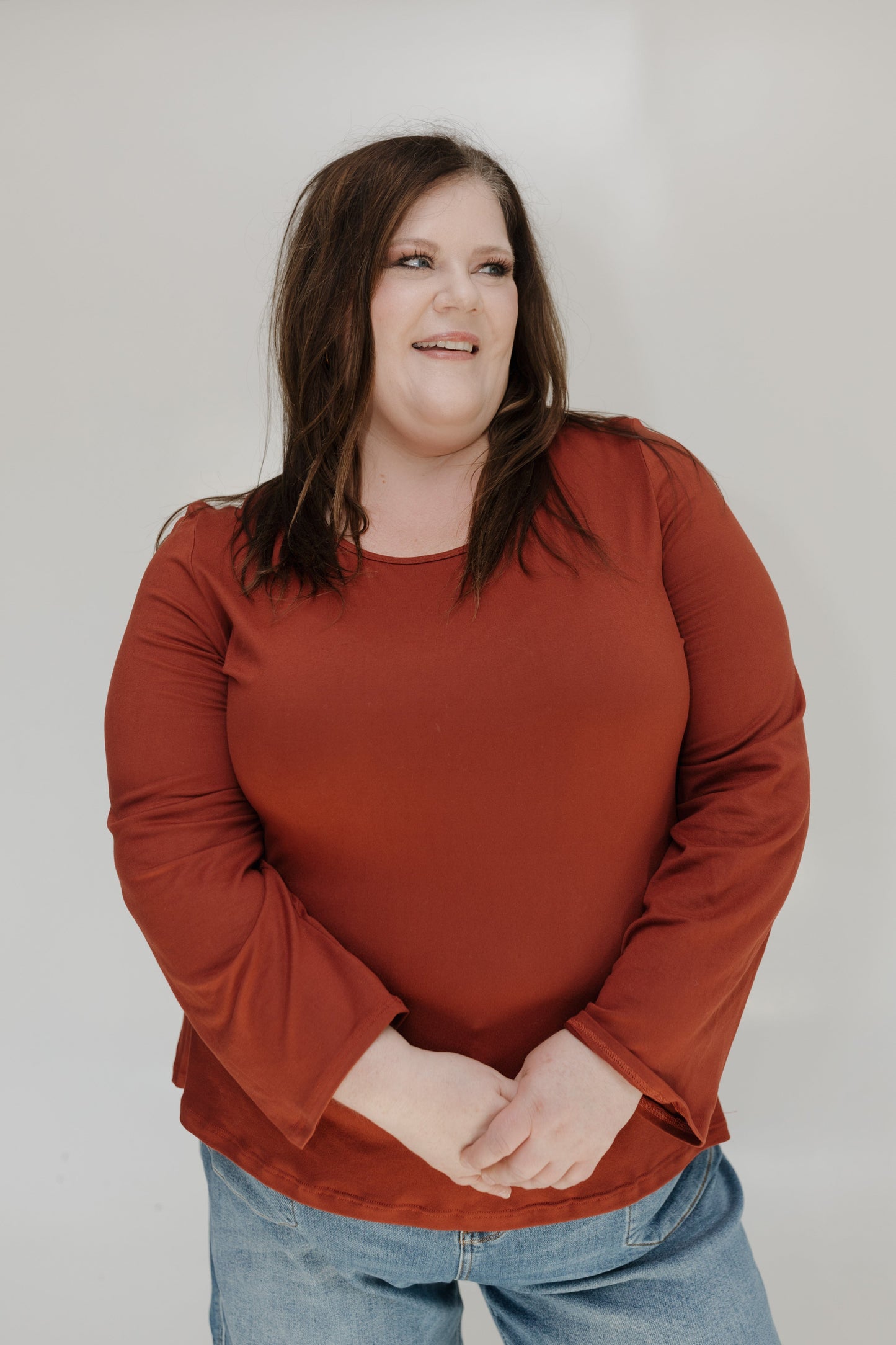 Woman wearing a rust-colored long-sleeve top and blue jeans against a plain background