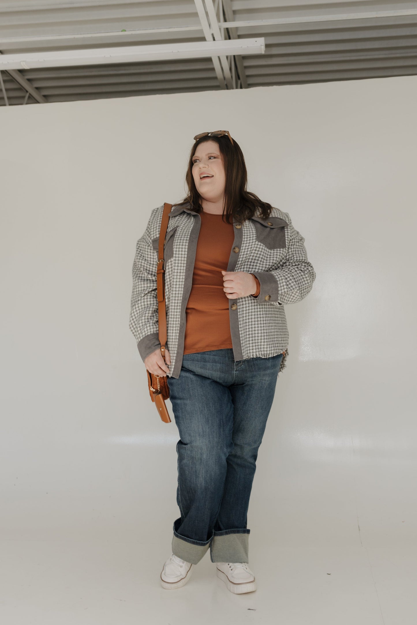 Woman wearing a plaid jacket, brown shirt, and jeans holding a brown bag against a white wall.