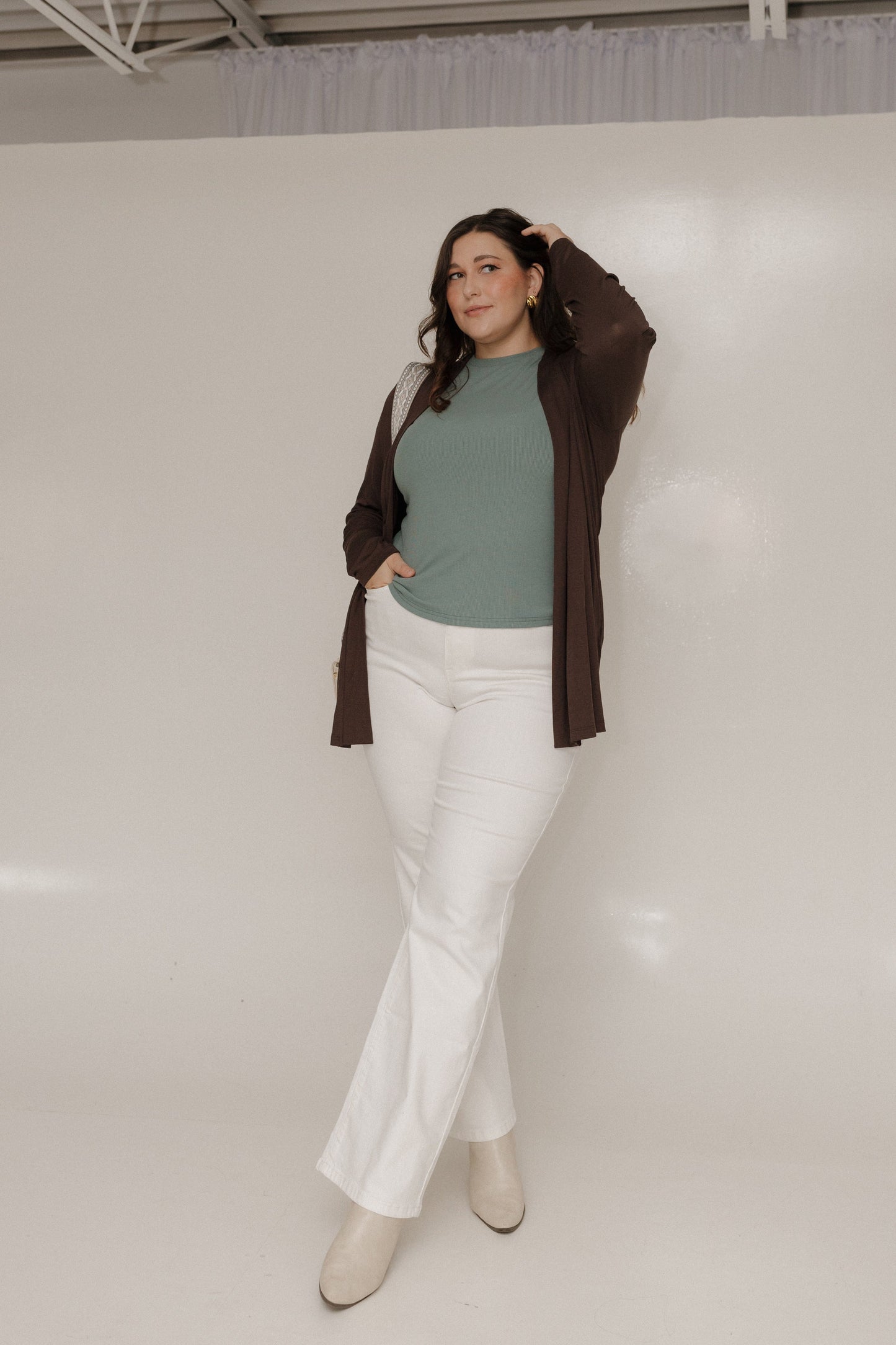 Woman wearing a green top, white pants, and a brown cardigan against a plain background