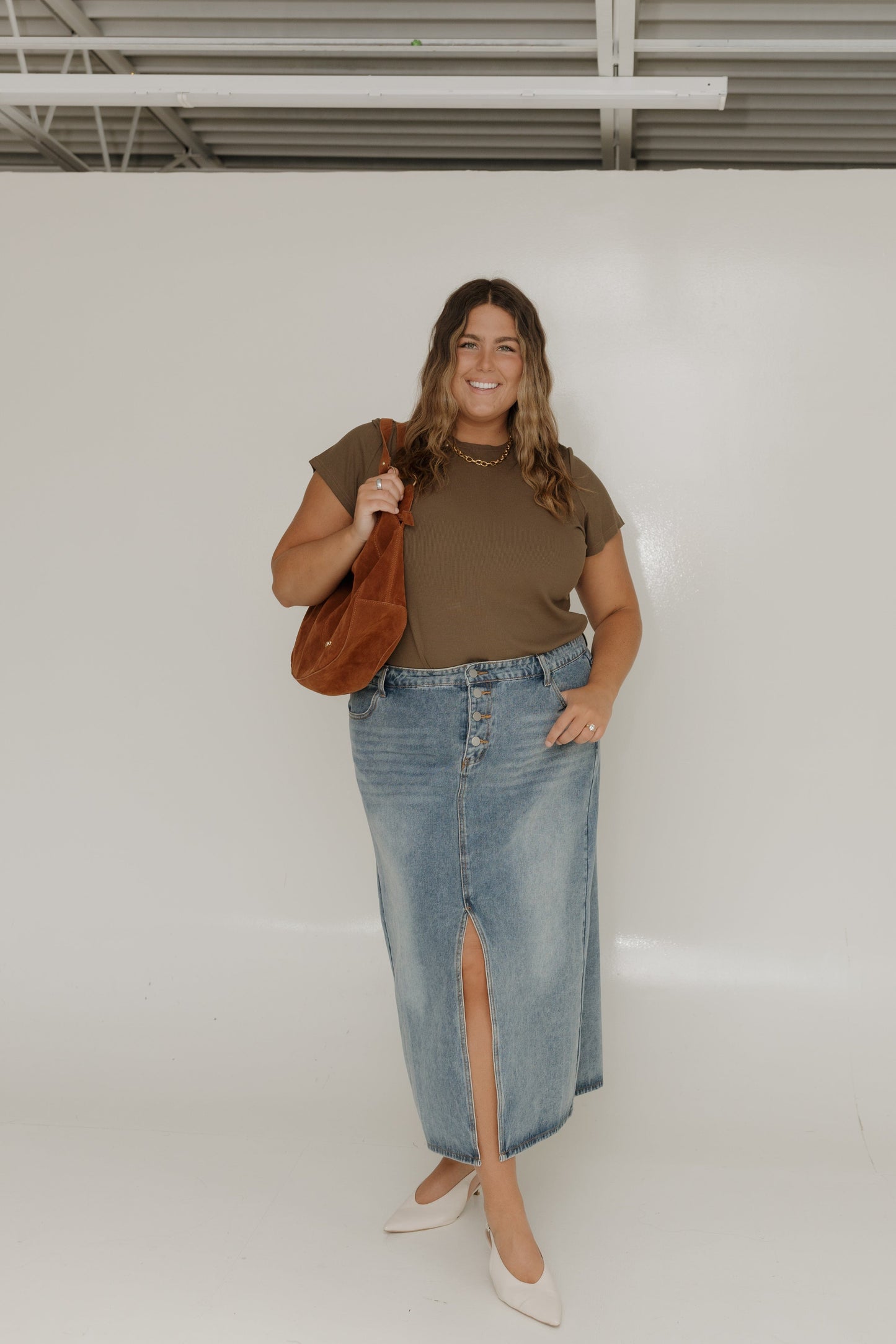 Woman wearing a brown top and blue denim skirt holding a brown bag against a white wall.
