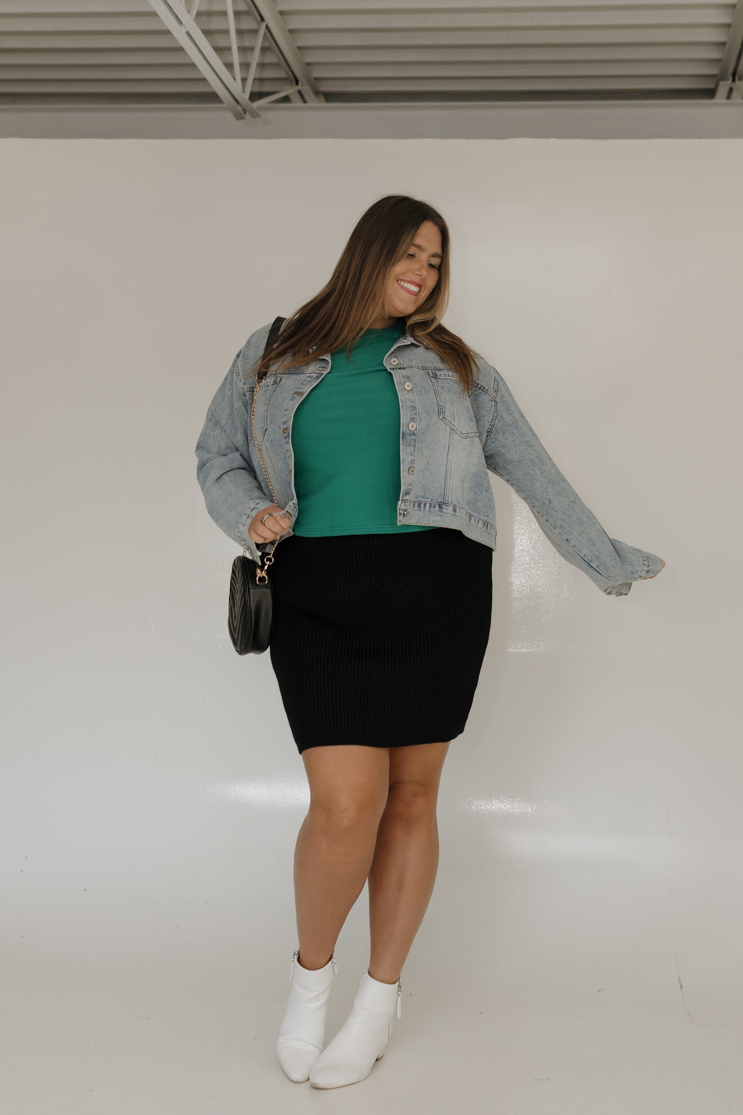 Woman wearing a denim jacket, green top, black skirt, and white boots against a white background