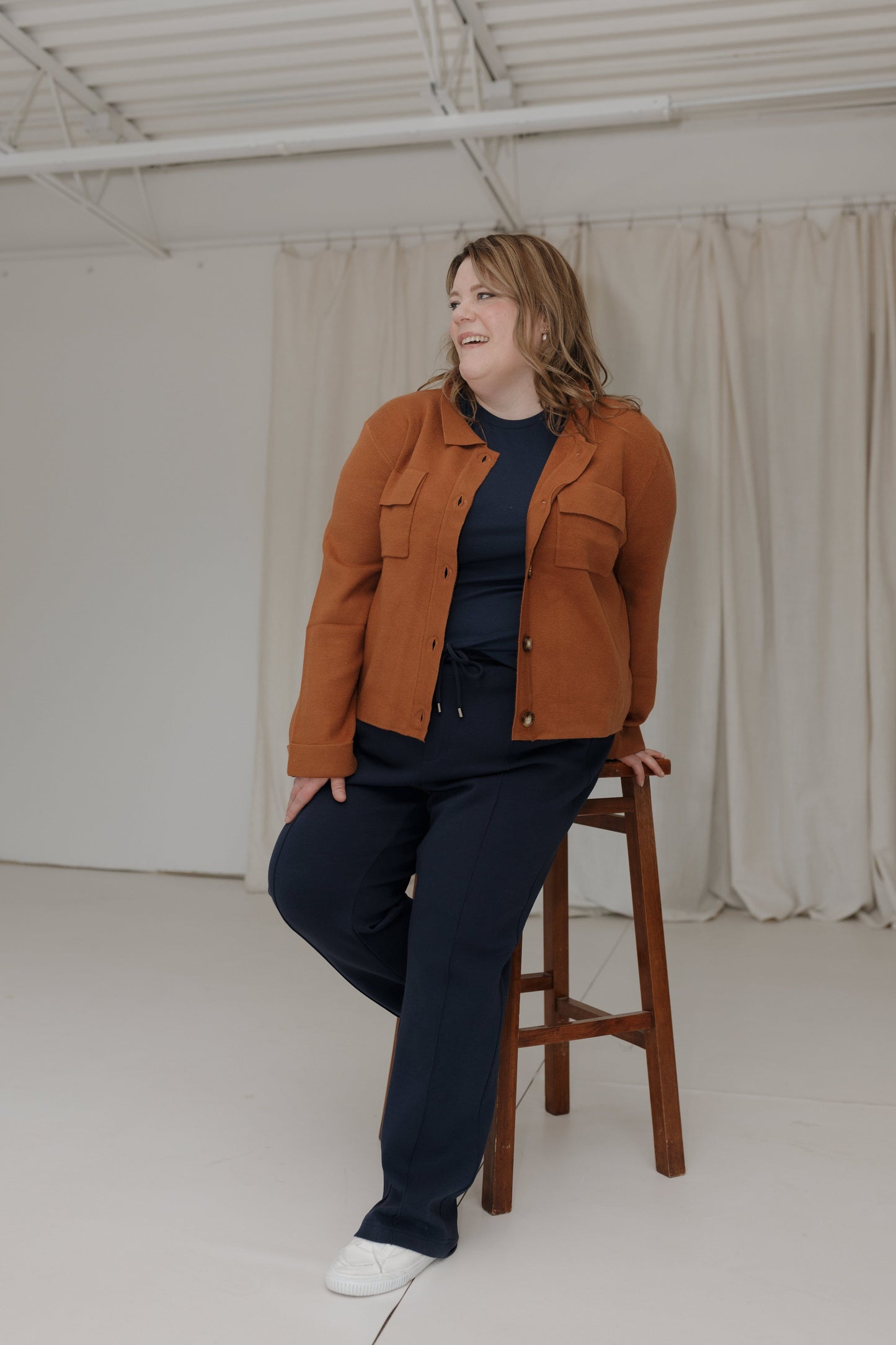 Person wearing a brown jacket and dark pants sitting on a wooden stool in a minimalistic room.