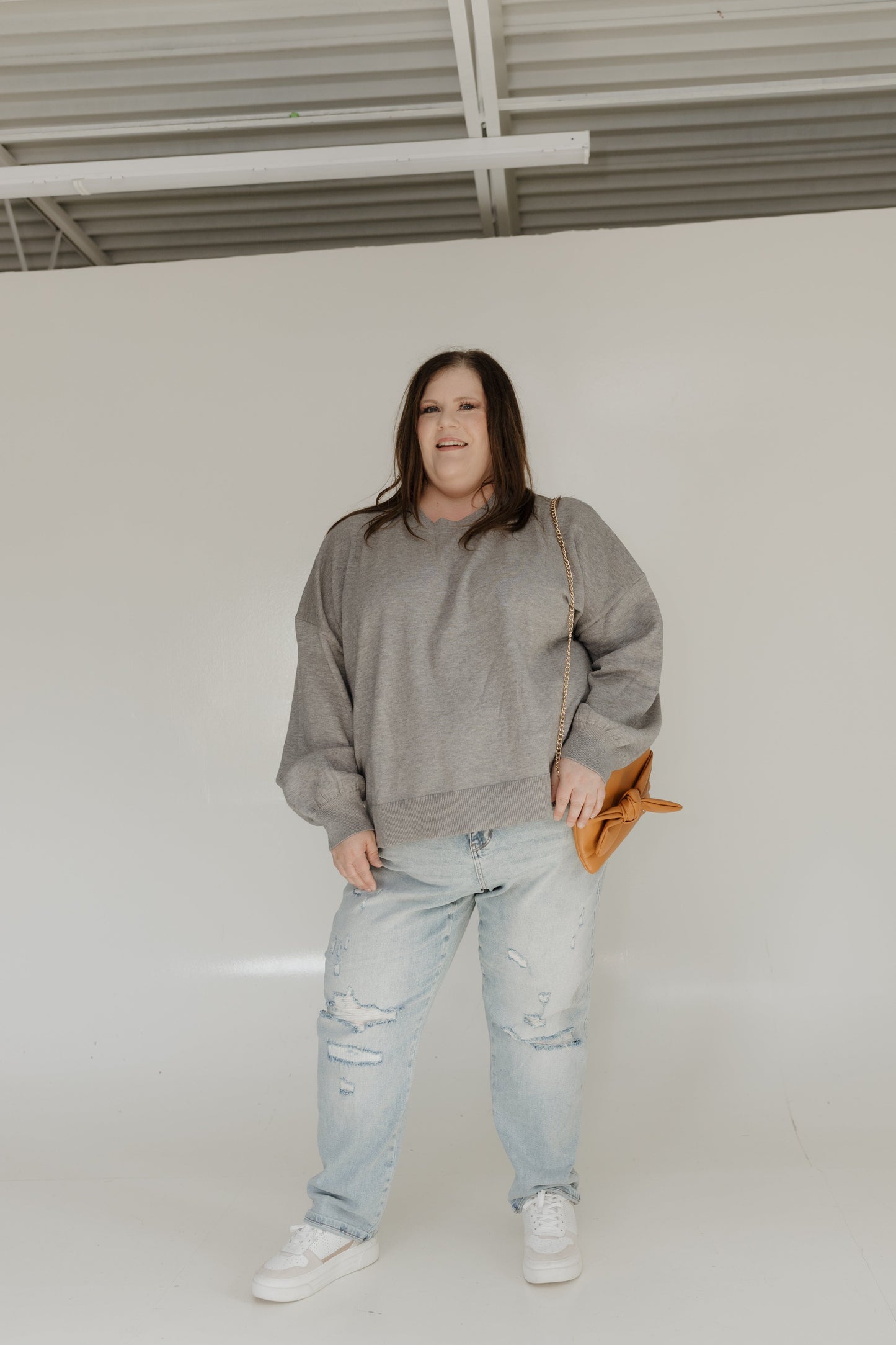Person wearing a gray sweatshirt and light blue jeans standing against a white wall.