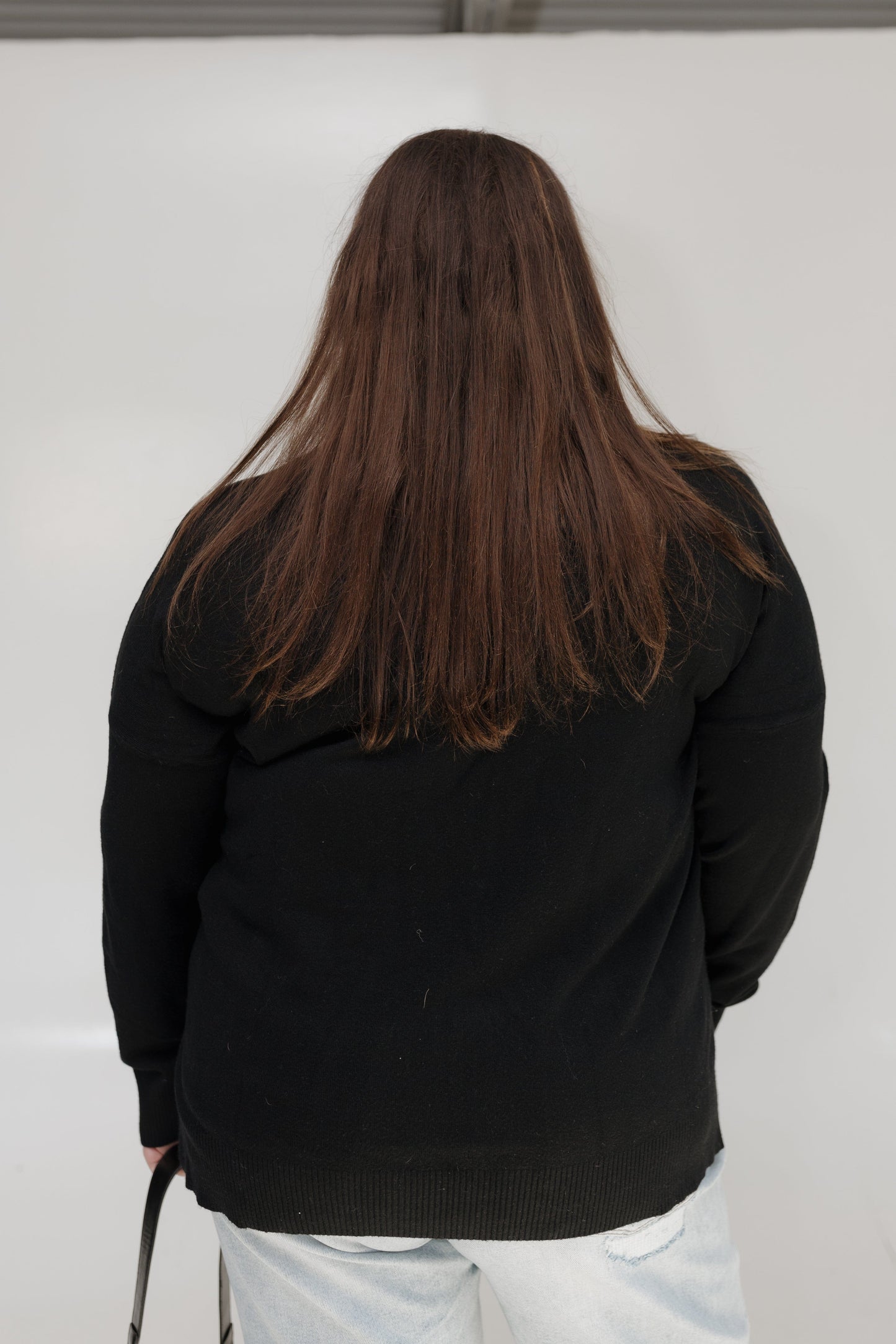 Person wearing a black jacket with long hair, standing against a white wall.