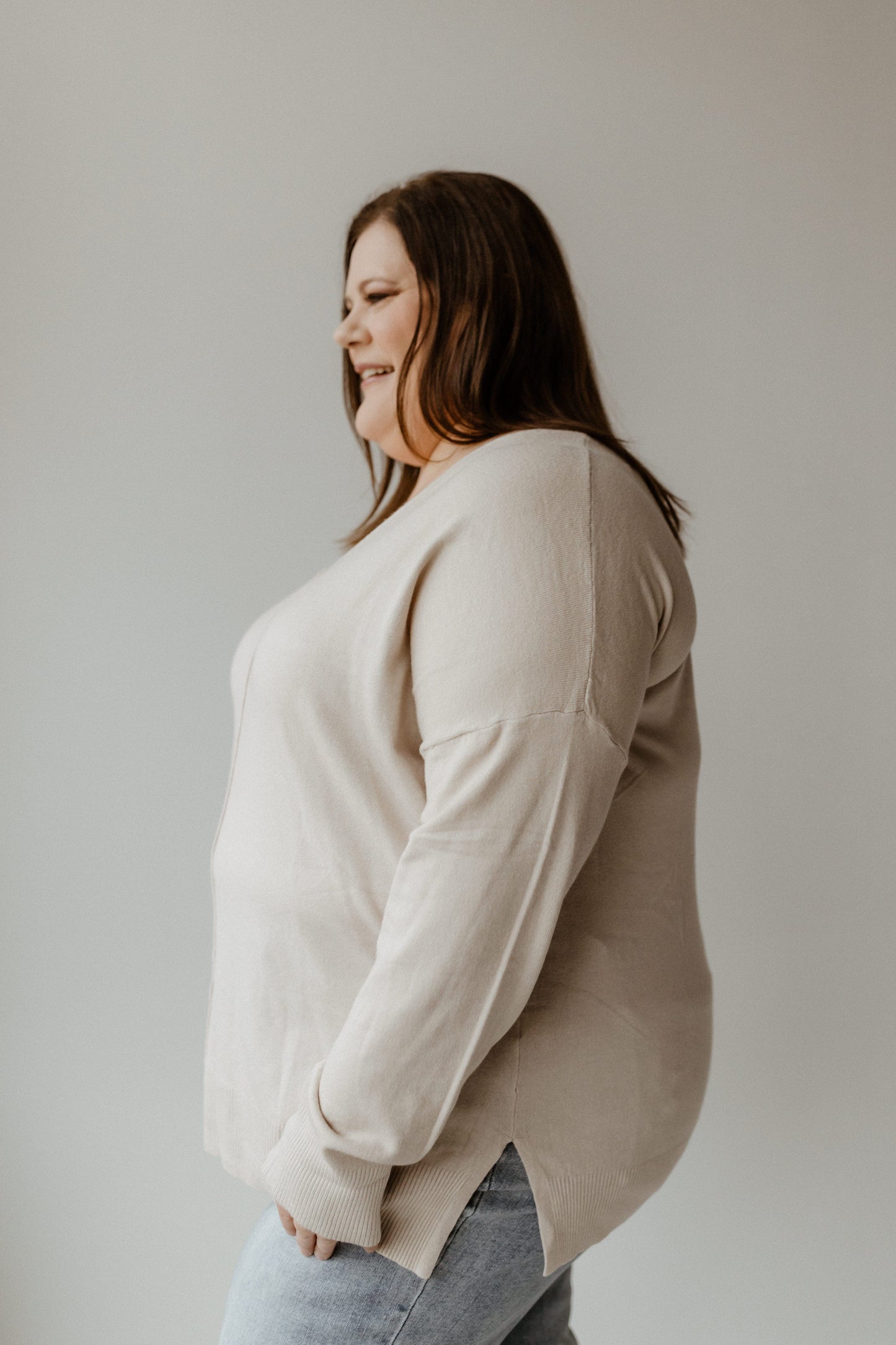 A person with long hair is smiling, standing sideways against a plain background, wearing a cozy V-Neck Front Seam Sweater and jeans.