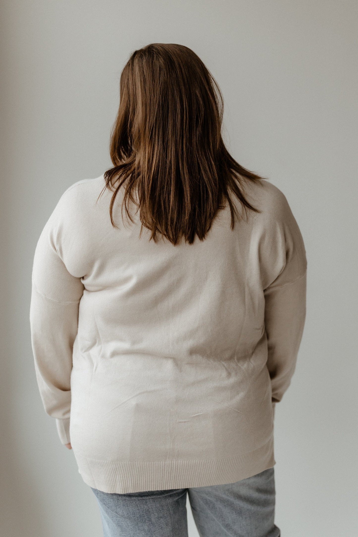 A person with long brown hair wearing a cozy beige V-Neck Front Seam Sweater and jeans stands facing away.