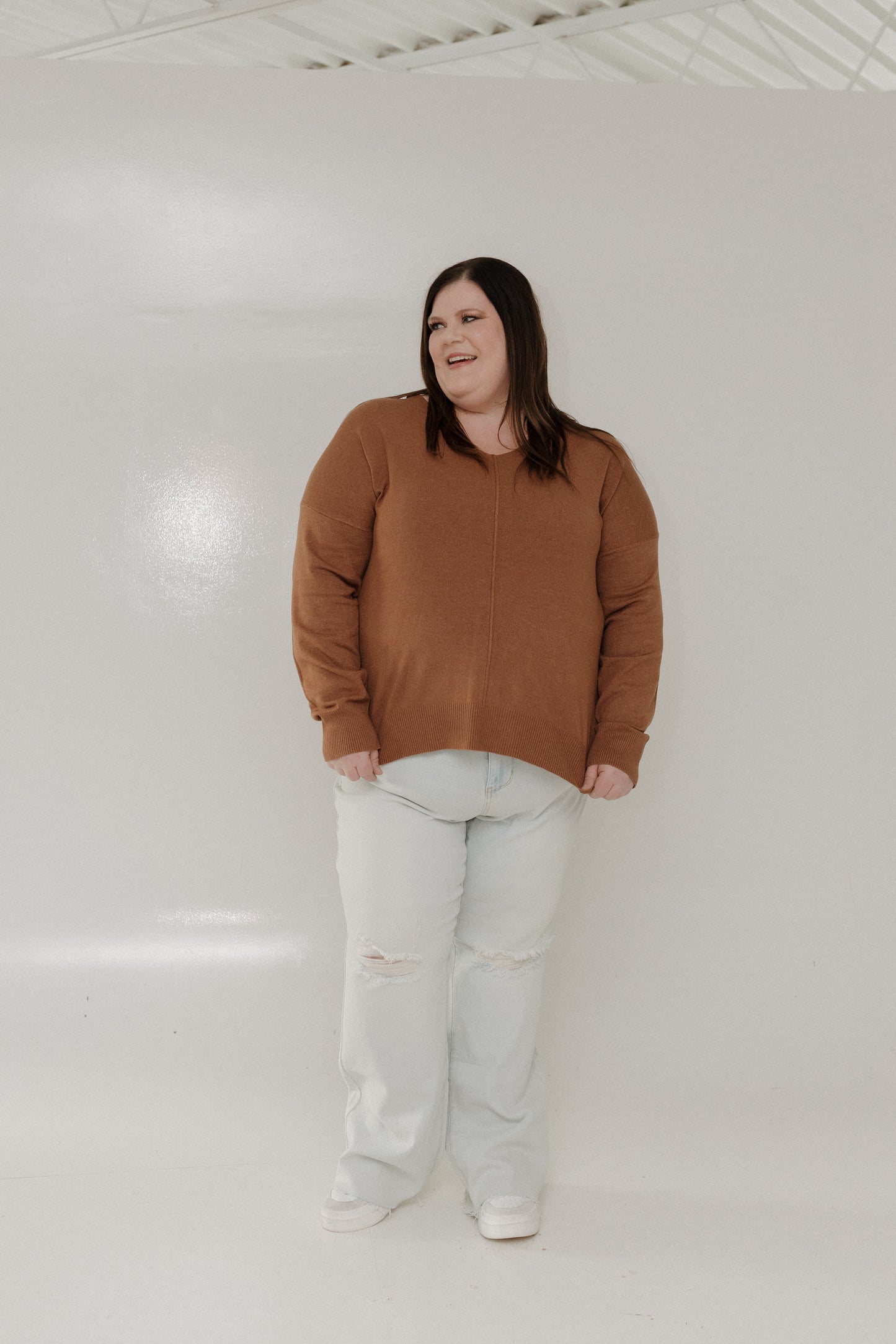 Person wearing a brown sweater and light-colored pants standing against a white wall.