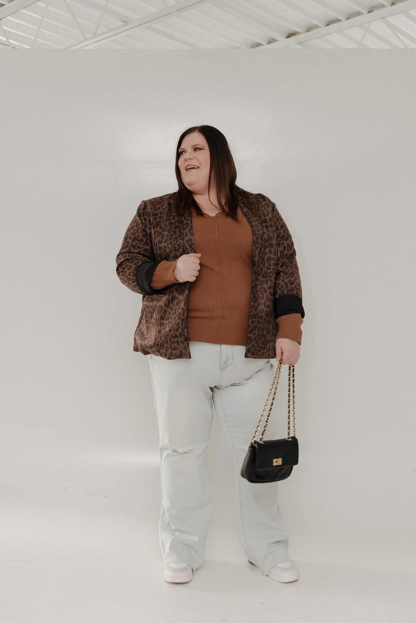 Woman wearing a leopard print jacket, brown top, light blue jeans, and holding a black handbag against a white background.
