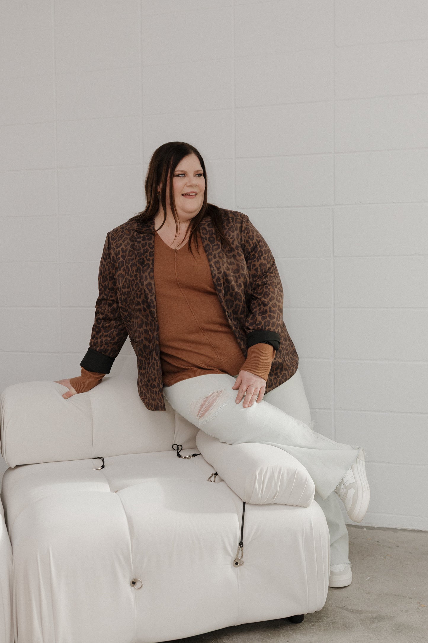 Woman sitting on a white chair wearing a brown jacket and white pants with a visible tear.