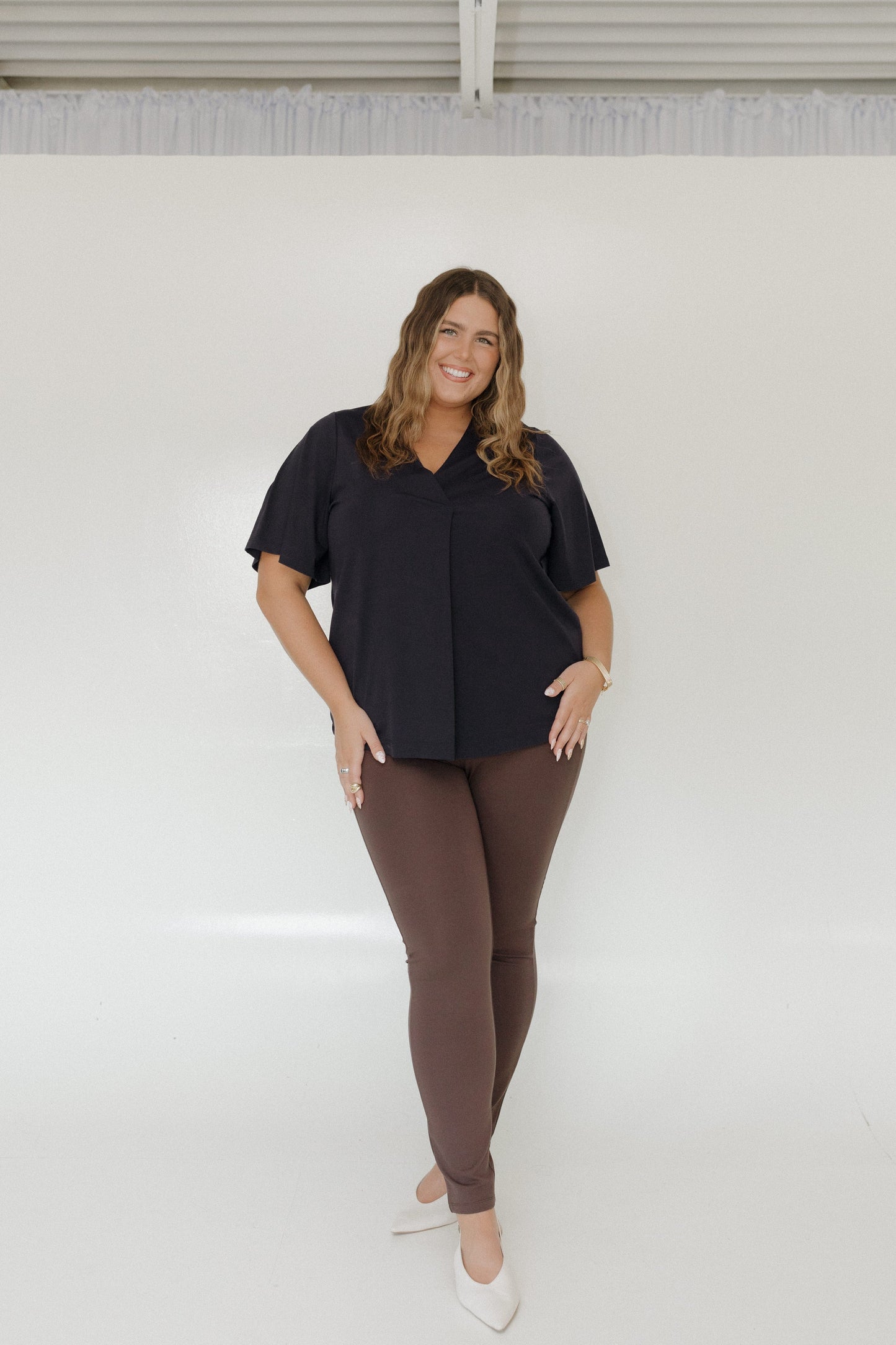 Woman wearing a black top and brown pants standing against a white wall.