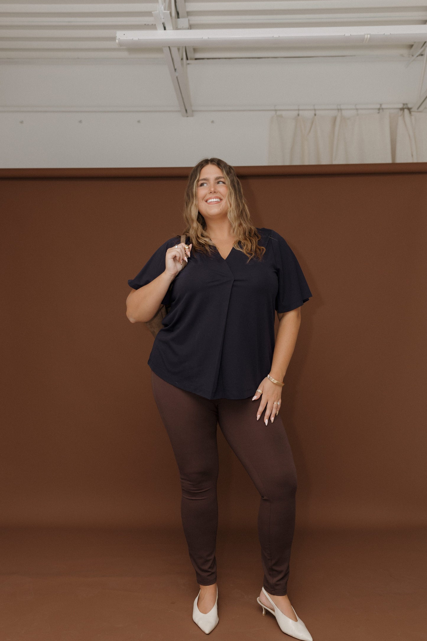 Woman wearing a black top and brown pants against a brown wall.