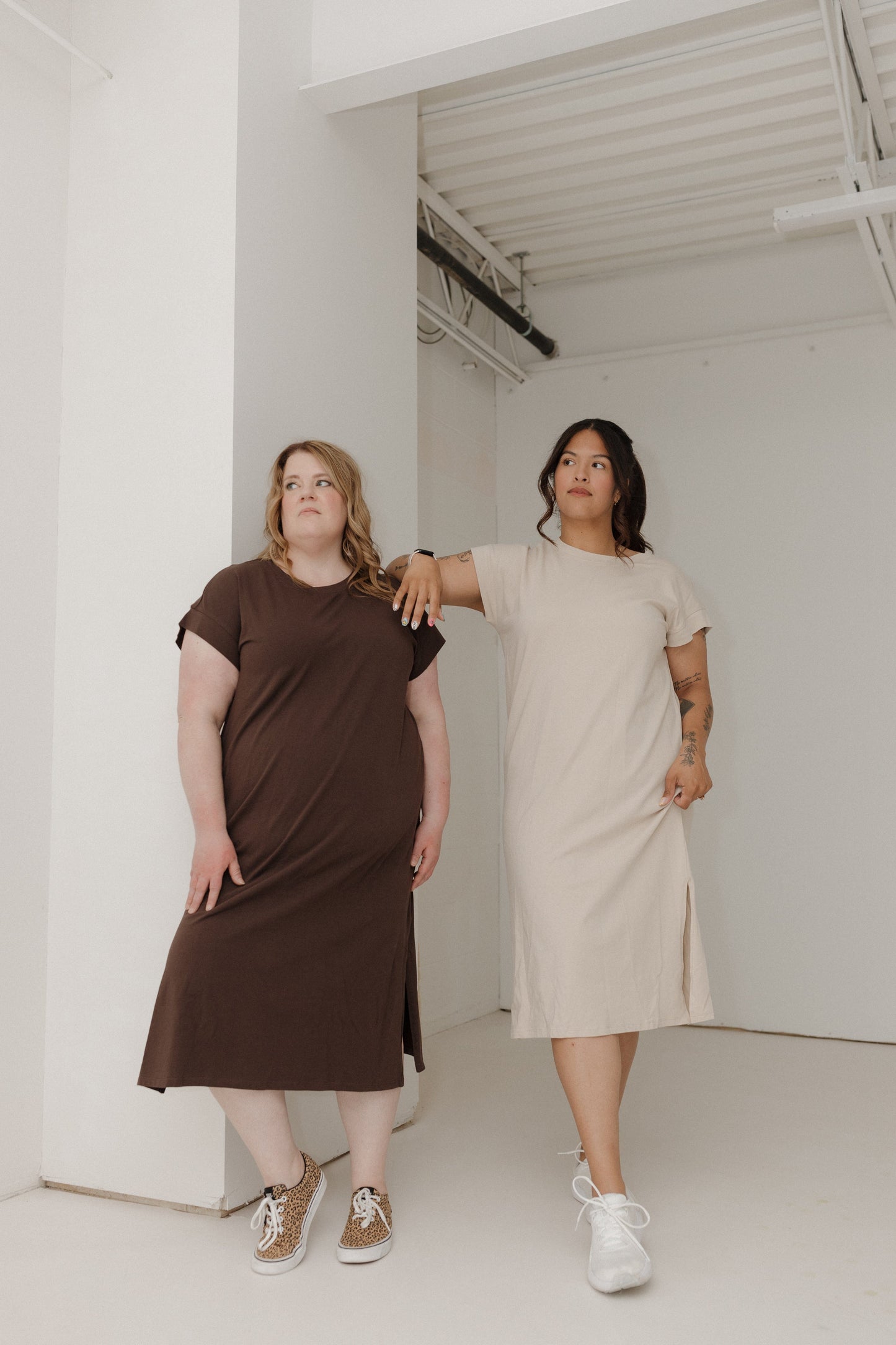 Two women standing in a minimal white room wearing brown and beige dresses.