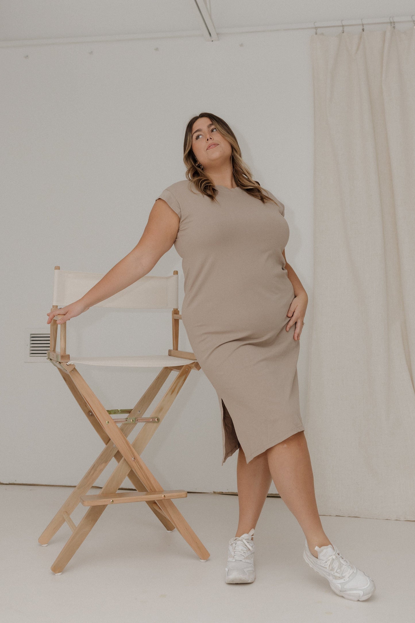 Woman in a beige dress standing next to a director's chair against a white wall.