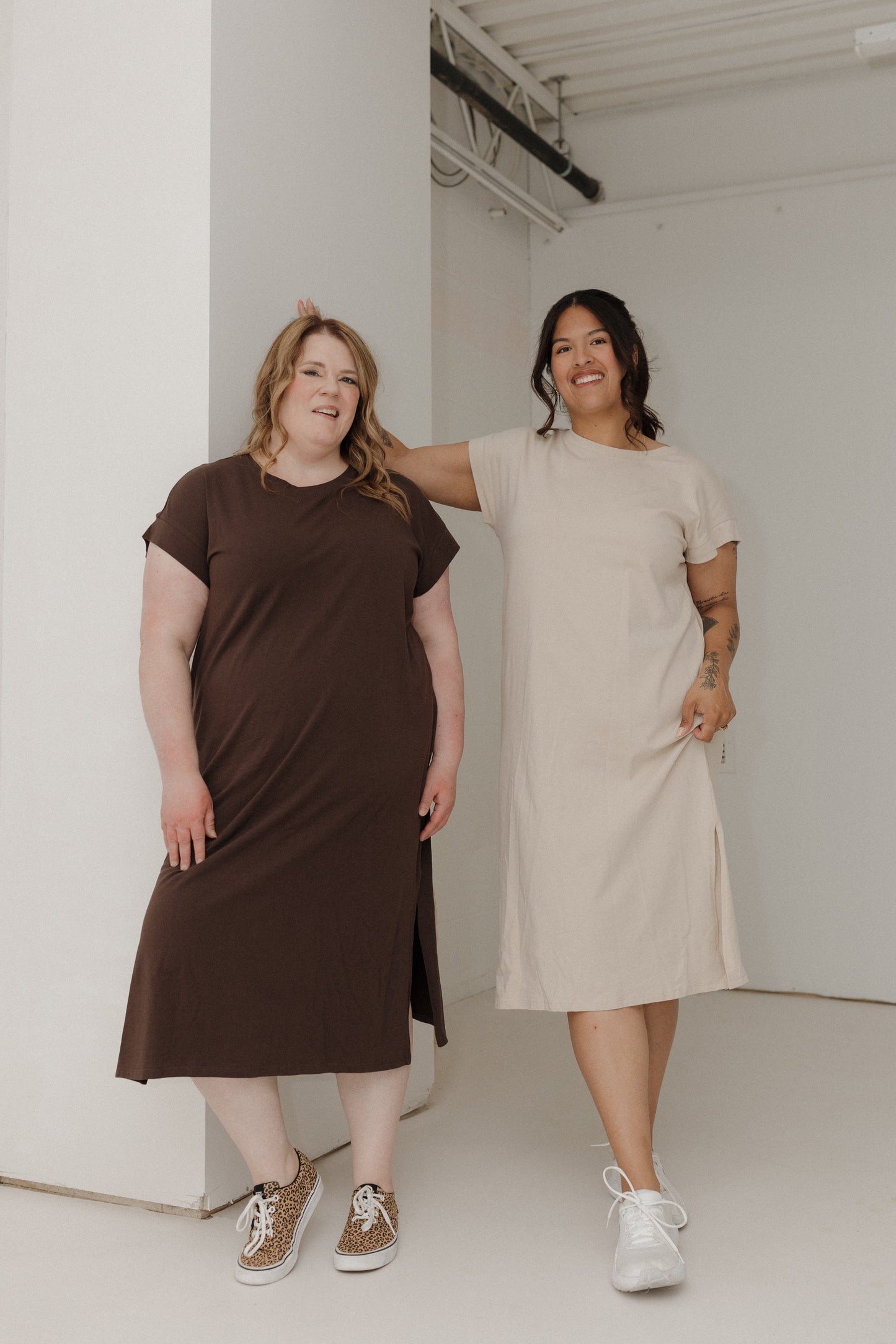 Two women wearing brown and beige dresses standing against a white wall.