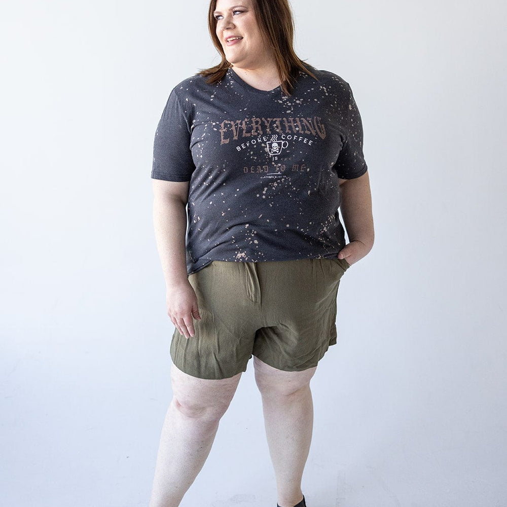 A person wearing the "EVERYTHING BEFORE COFFEE IS DEAD TO ME" graphic tee by Love Marlow and green shorts, standing against a plain white background.