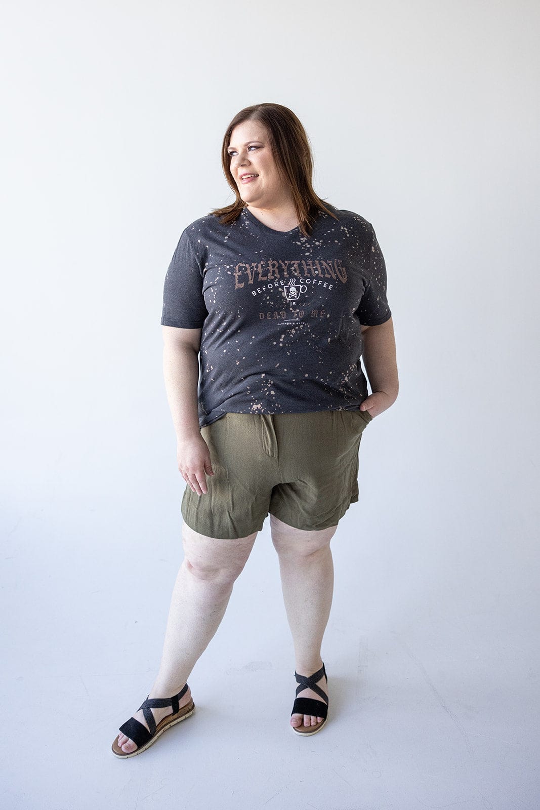 A person wearing the "EVERYTHING BEFORE COFFEE IS DEAD TO ME" graphic tee by Love Marlow and green shorts, standing against a plain white background.