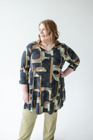 A person with long light brown hair is standing and smiling, wearing the EXTRA LONG TUNIC WITH V-NECK IN GEOMETRIC PATTERN by Love Marlow and beige pants, set against a plain white background.