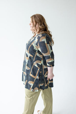 A woman with long wavy hair is standing sideways, wearing the Love Marlow EXTRA LONG TUNIC WITH V-NECK IN GEOMETRIC PATTERN and light-colored pants. She is set against a plain, light background.