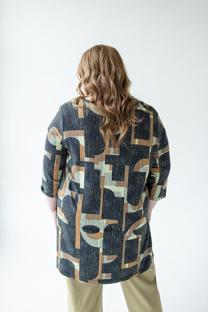 A person with wavy, blonde hair is standing with their back to the camera, wearing the Love Marlow EXTRA LONG TUNIC WITH V-NECK IN GEOMETRIC PATTERN and khaki pants.