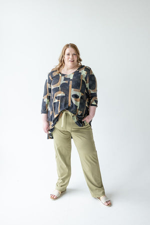 A person stands in front of a plain background, wearing an EXTRA LONG TUNIC WITH V-NECK IN GEOMETRIC PATTERN by Love Marlow, paired with khaki pants and sandals.