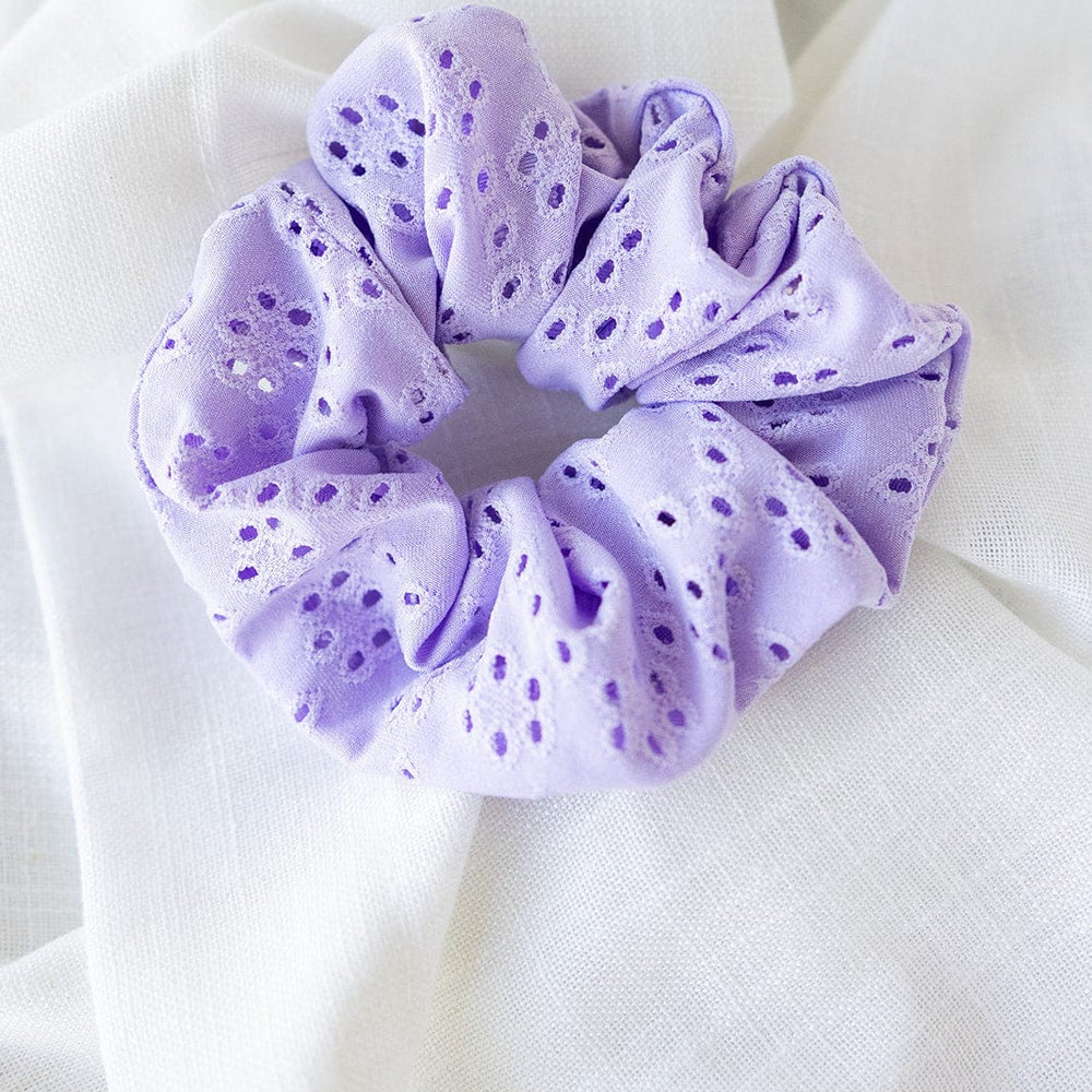 A lilac eyelet lace scrunchie rests gracefully on a white fabric background, showcasing the charm of handmade hair accessories.