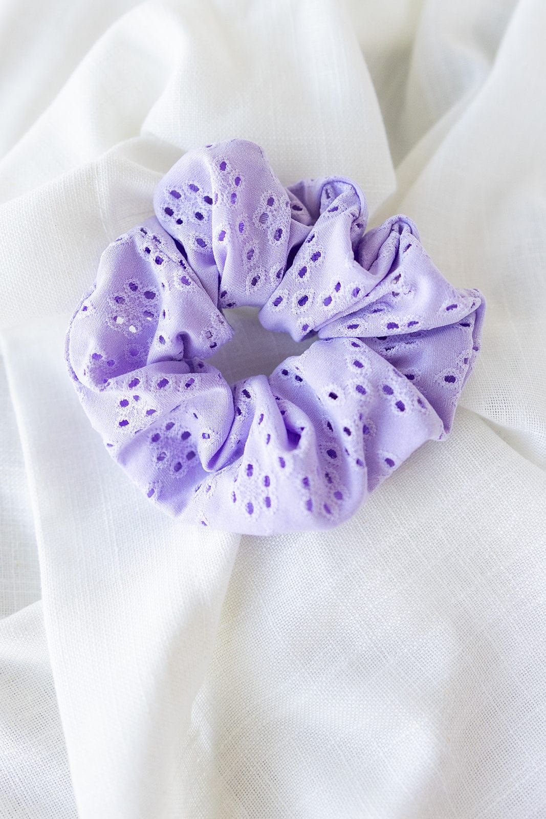 A lilac eyelet lace scrunchie rests gracefully on a white fabric background, showcasing the charm of handmade hair accessories.