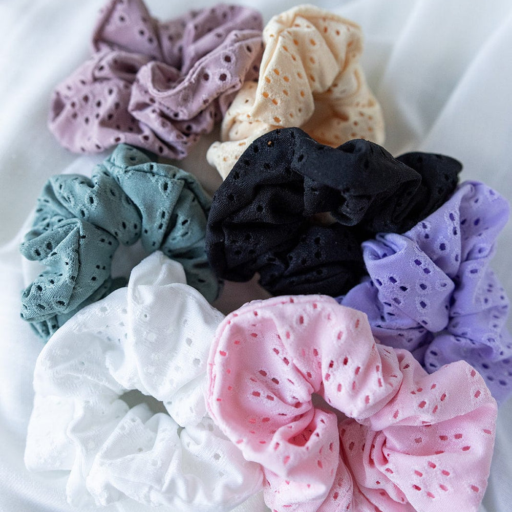 A collection of handmade hair accessories, featuring fabric scrunchies in an array of colors such as pink, white, black, purple, green, beige, and mauve. These delicate pieces include a charming lilac scrunchie displayed elegantly on a soft white surface.