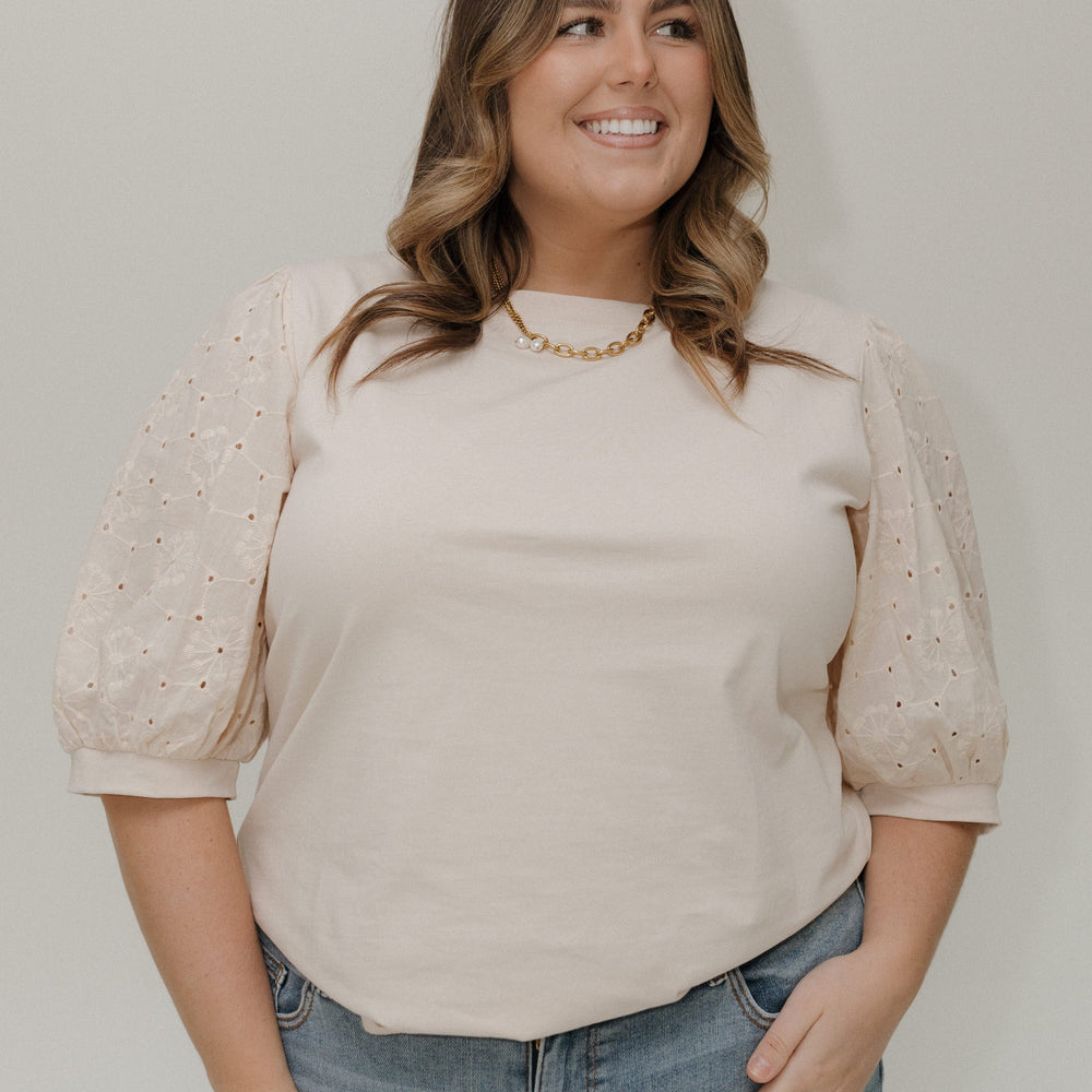Woman wearing a beige blouse with lace sleeves and blue jeans against a plain background