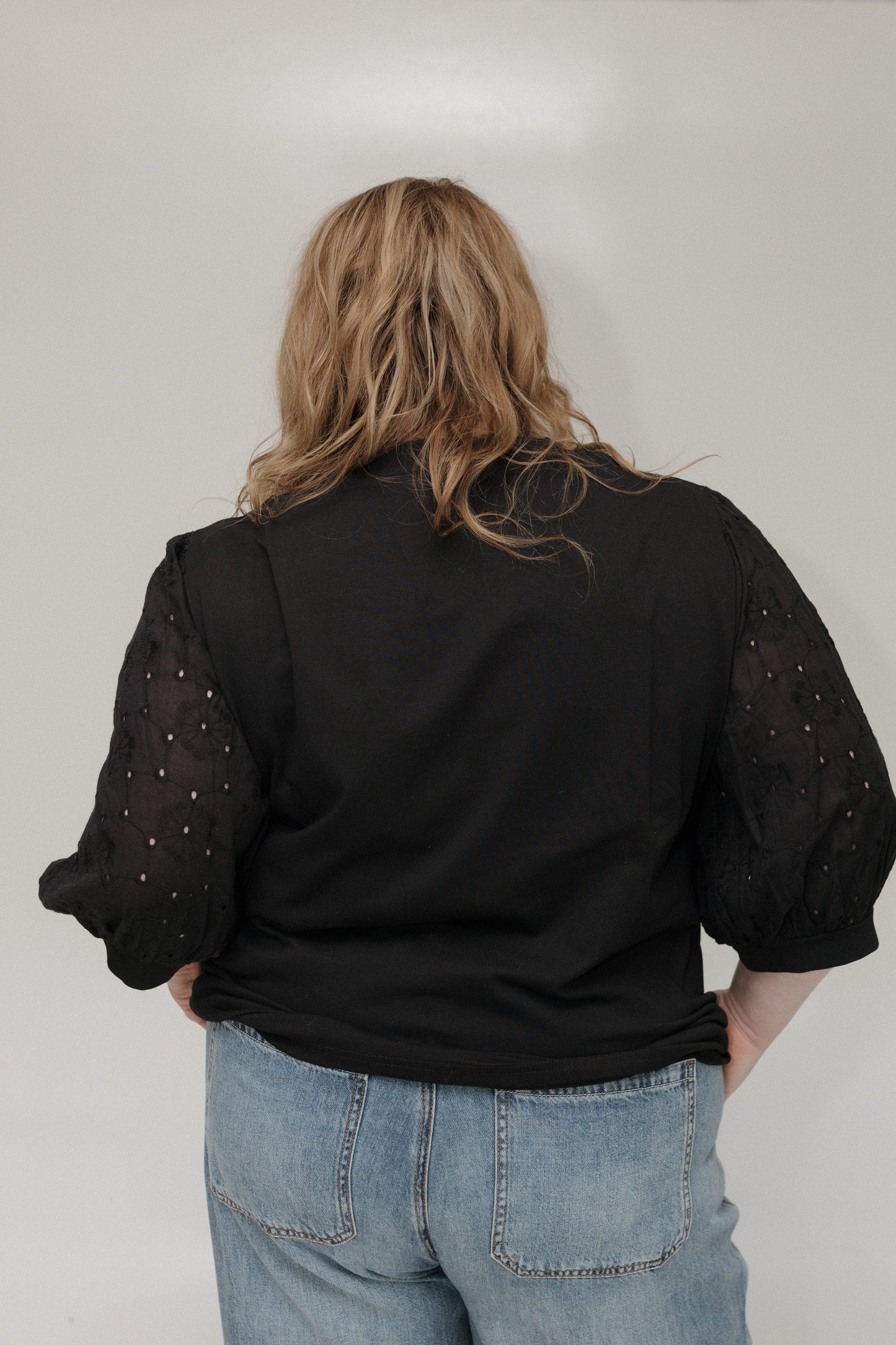 Person wearing a black top with embellished sleeves and light blue jeans against a plain background