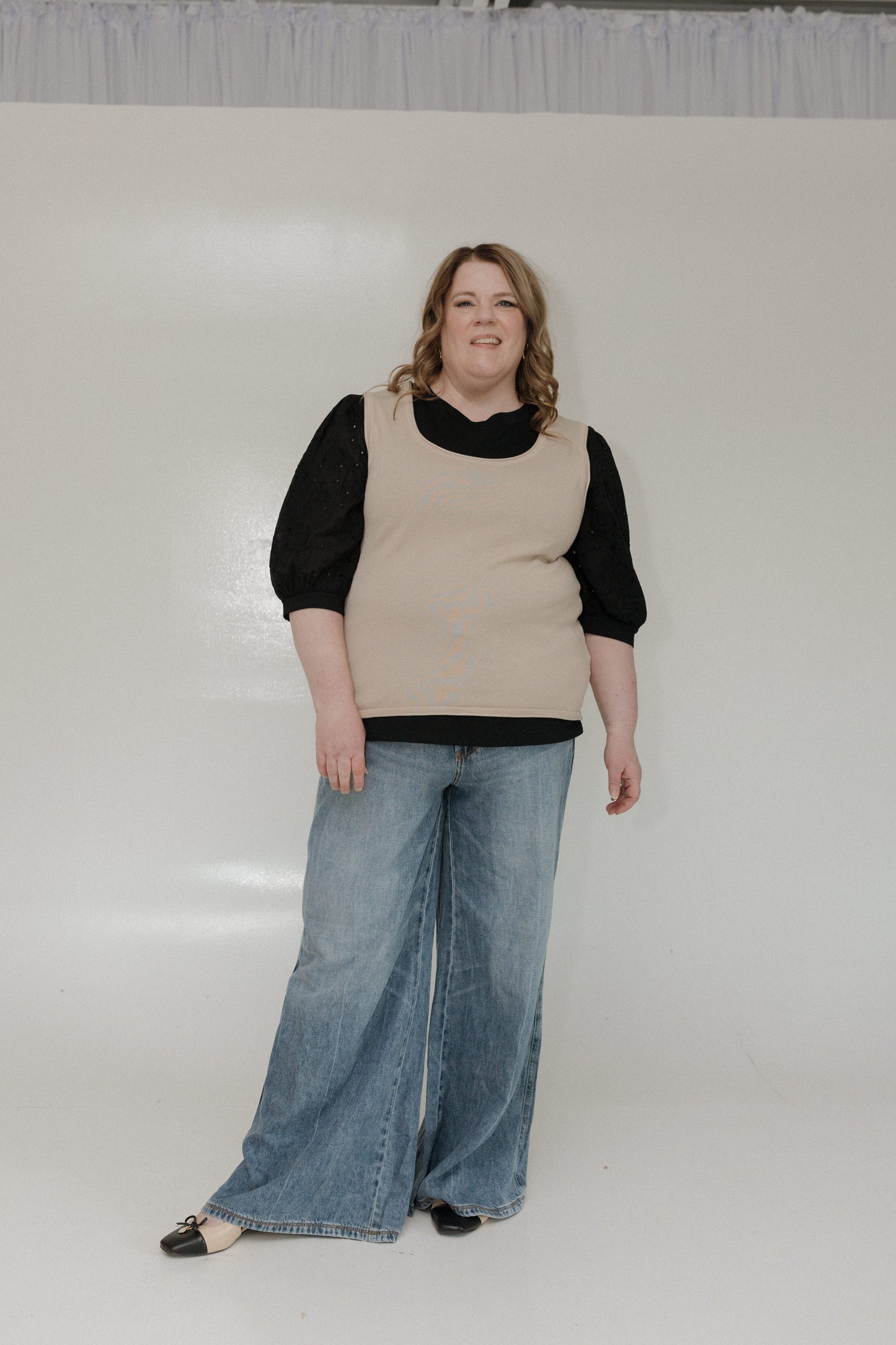 Person wearing a beige vest over a black shirt and blue jeans on a white background