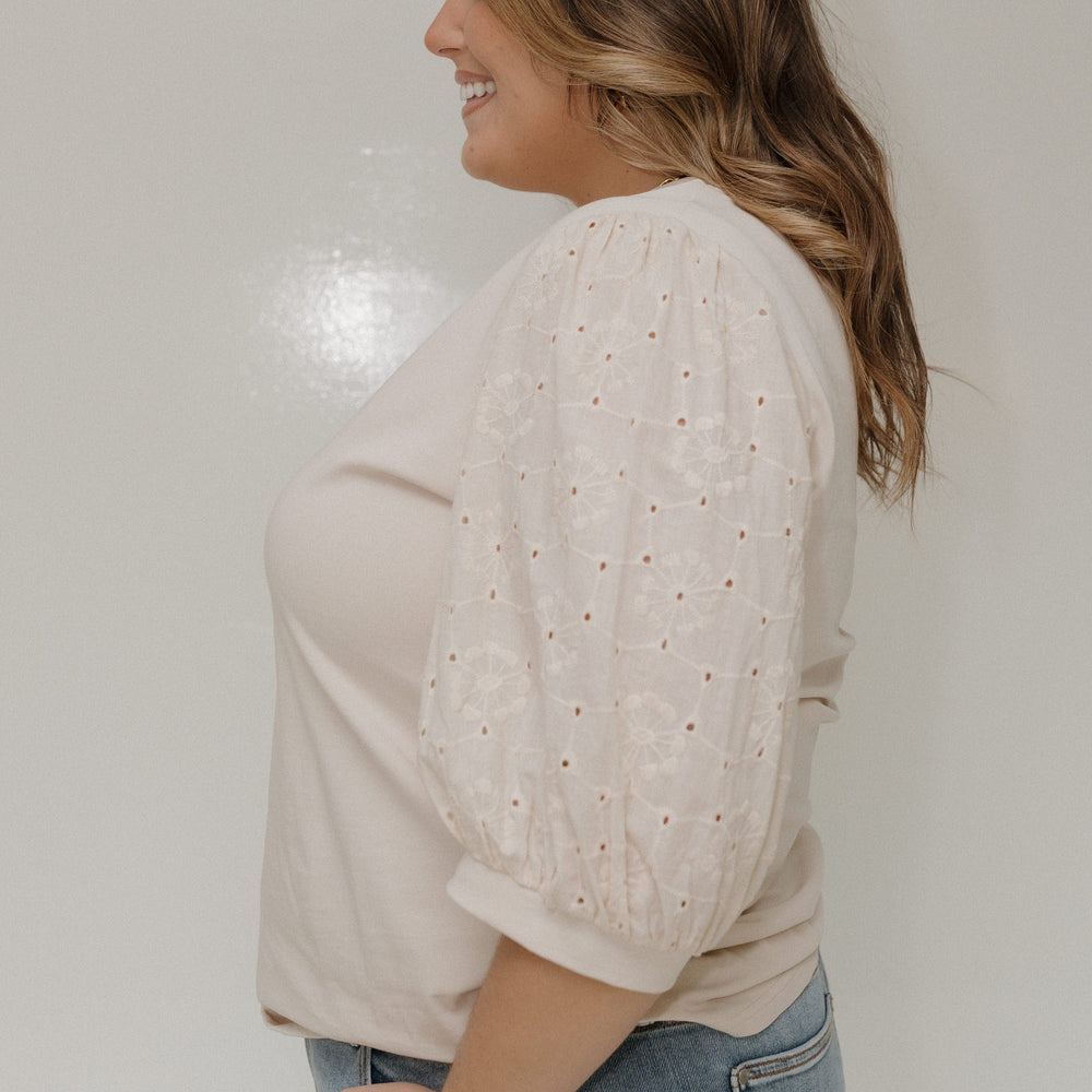 Woman wearing a beige lace top and jeans against a plain background