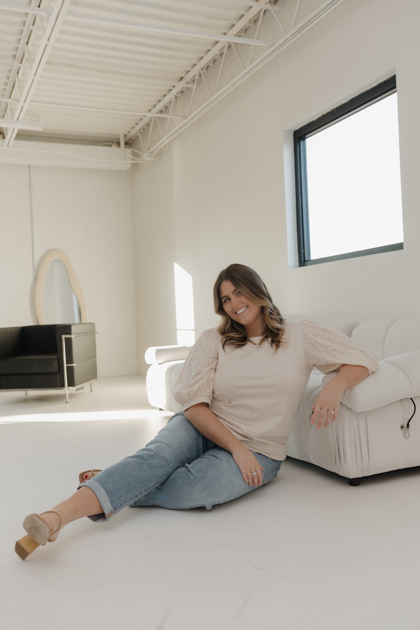 Woman sitting on a white couch in a minimalistic room with a large window.