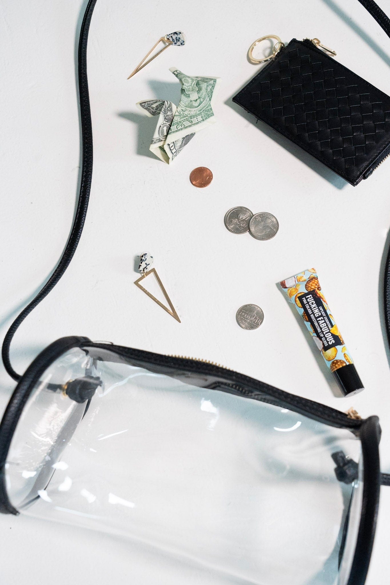 Clear pouch with black strap, wallet, coins, earrings, and other small items on a white background