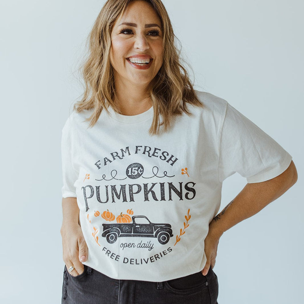 A woman is smiling, wearing the ultra-soft "Farm Fresh Pumpkins" Graphic Tee in Ivory by Love Marlow, featuring a charming graphic of a truck filled with pumpkins that captures the spirit of the autumn harvest. She stands against a plain light background with her hands in her jeans pockets.
