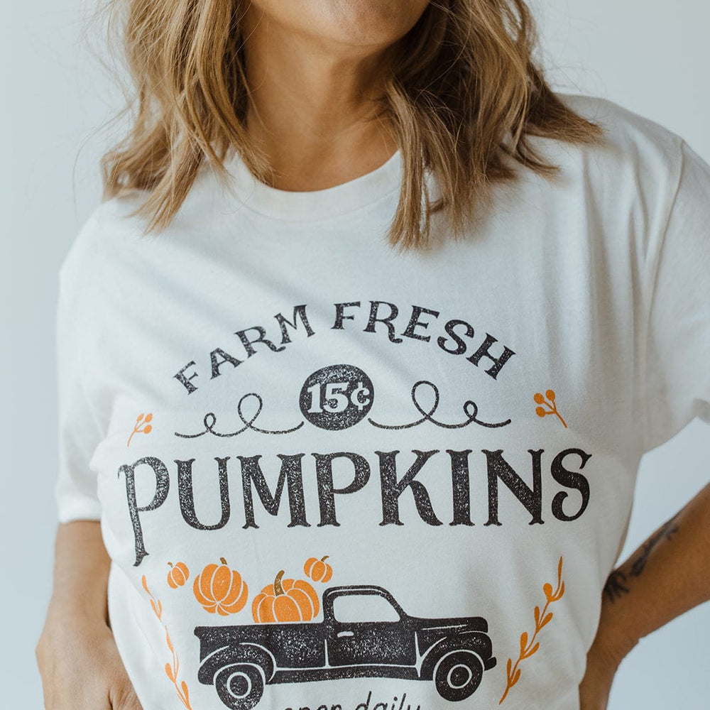 A person wearing Love Marlow's ultra-soft "Farm Fresh Pumpkins" Graphic Tee in Ivory, featuring a charming truck and pumpkins design, alongside text that reads "15¢," "open daily," and "Free Deliveries," perfectly capturing the essence of the autumn harvest.