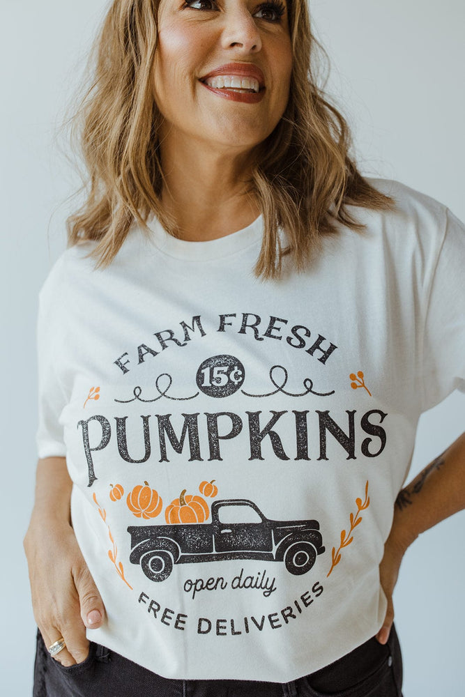 A person wearing Love Marlow's ultra-soft "Farm Fresh Pumpkins" Graphic Tee in Ivory, featuring a charming truck and pumpkins design, alongside text that reads "15¢," "open daily," and "Free Deliveries," perfectly capturing the essence of the autumn harvest.