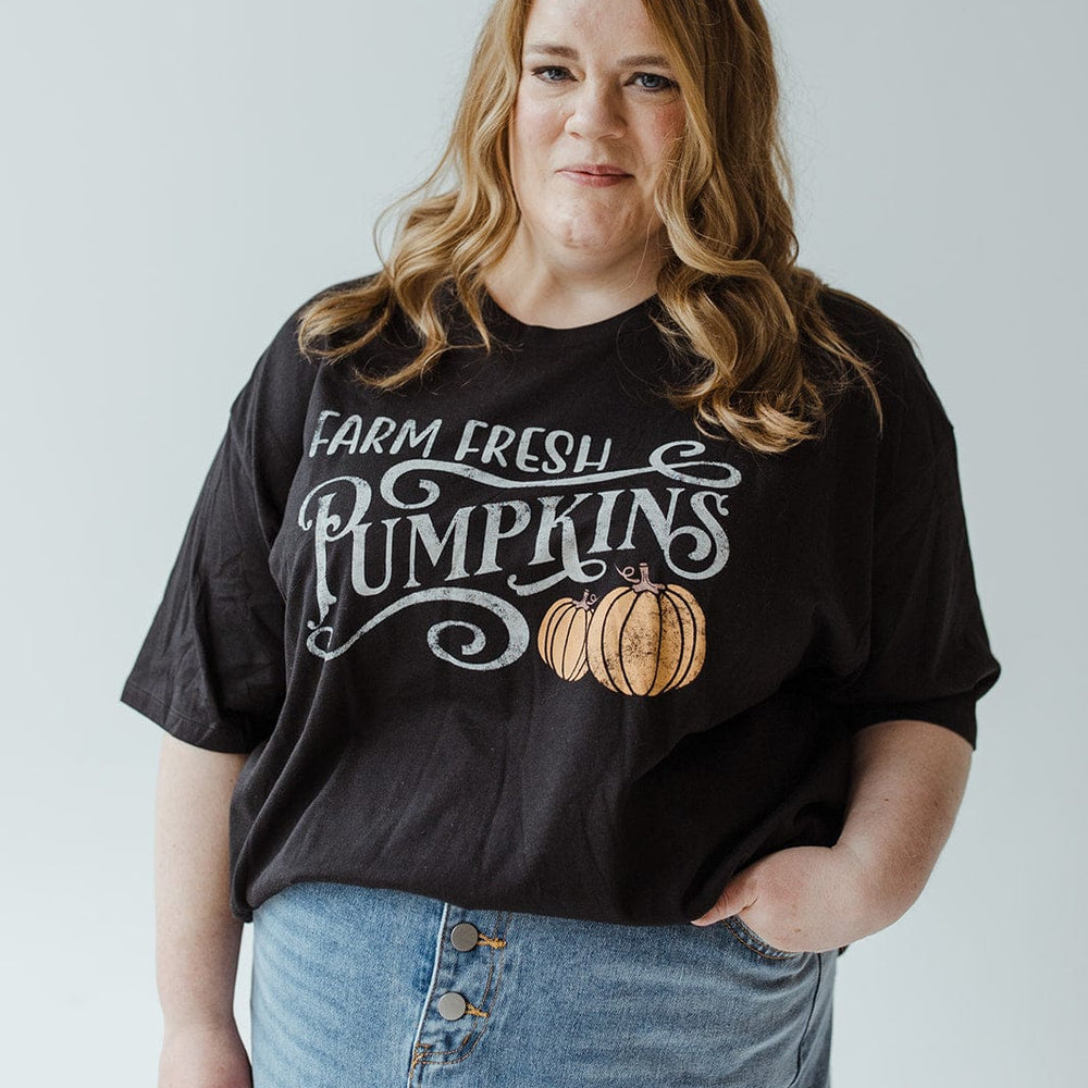 A person with wavy hair wears the "FARM FRESH PUMPKINS" graphic tee in black from Love Marlow, featuring super-soft fabric, paired with a denim skirt while standing against a plain background.