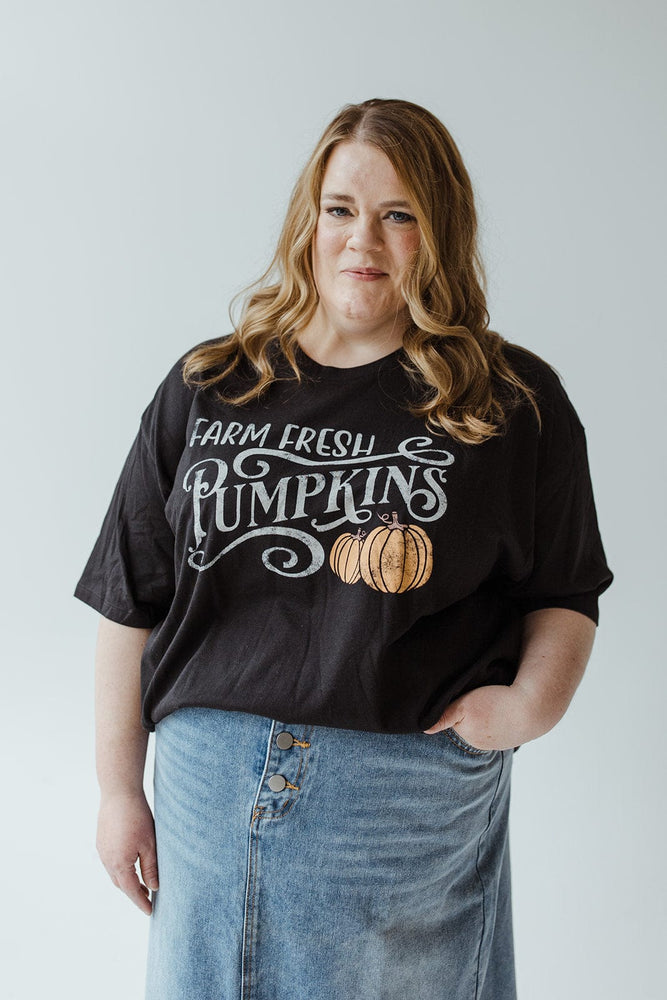 A person with wavy hair wears the "FARM FRESH PUMPKINS" graphic tee in black from Love Marlow, featuring super-soft fabric, paired with a denim skirt while standing against a plain background.