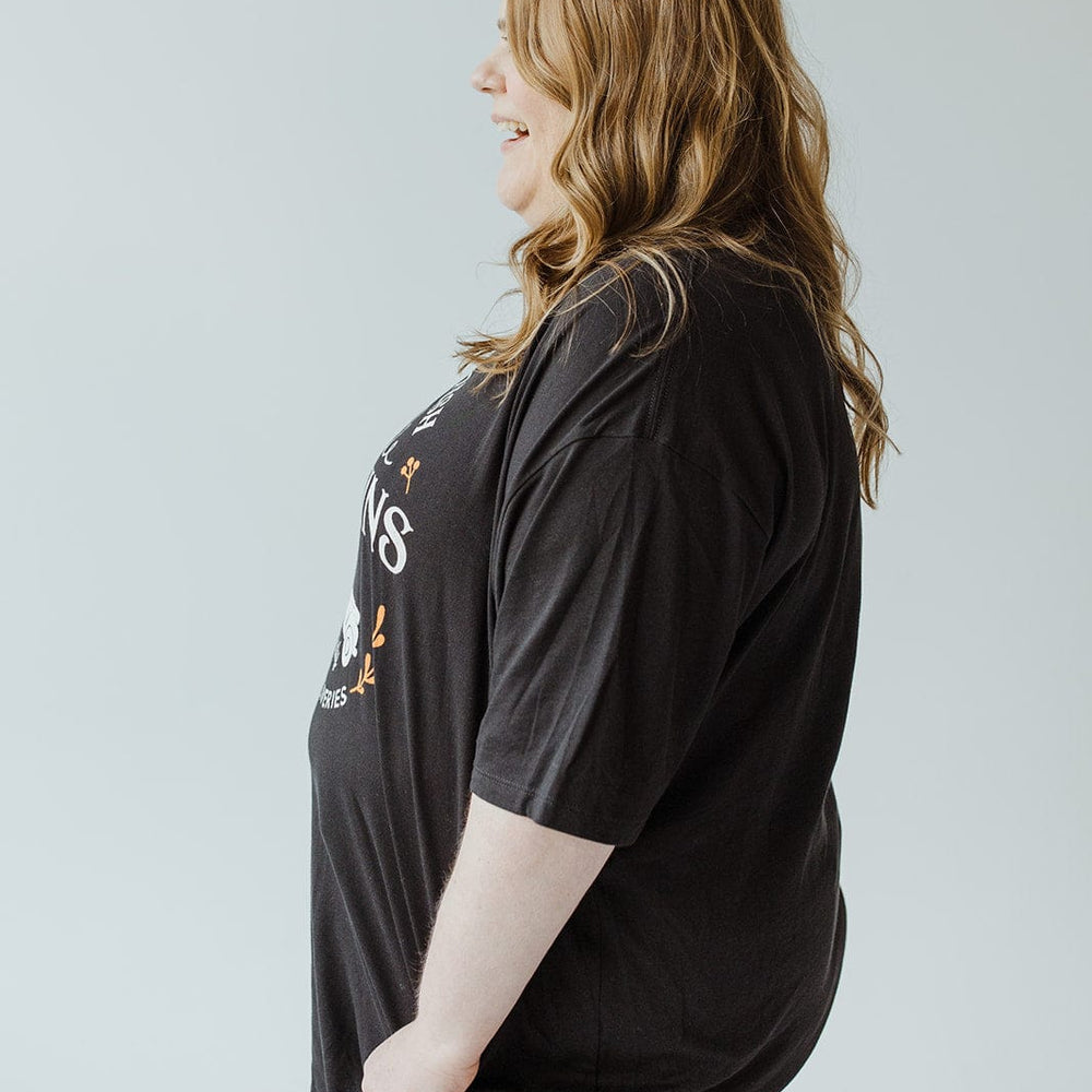 A smiling woman with long hair stands in profile, wearing the "FARM FRESH PUMPKINS" with truck graphic tee in black by Love Marlow, paired with jeans, against a plain background.