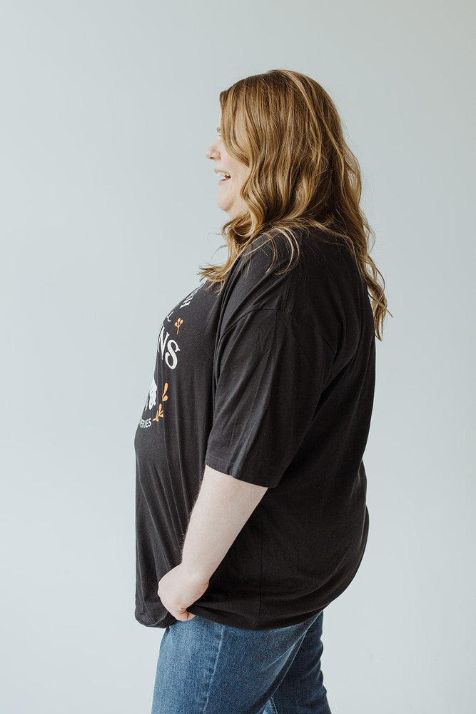 A smiling woman with long hair stands in profile, wearing the "FARM FRESH PUMPKINS" with truck graphic tee in black by Love Marlow, paired with jeans, against a plain background.