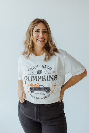 A woman is smiling, wearing the ultra-soft "Farm Fresh Pumpkins" Graphic Tee in Ivory by Love Marlow, featuring a charming graphic of a truck filled with pumpkins that captures the spirit of the autumn harvest. She stands against a plain light background with her hands in her jeans pockets.