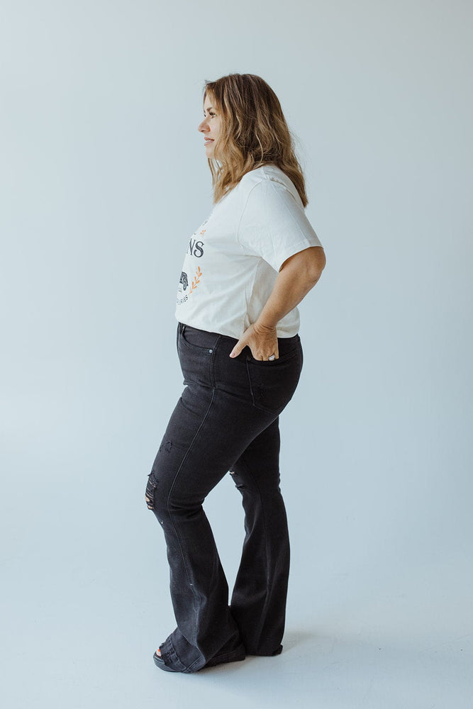 A woman with long hair stands in profile, wearing the "FARM FRESH PUMPKINS" graphic tee in ivory by Love Marlow, paired with dark jeans and black shoes, against a plain white background.