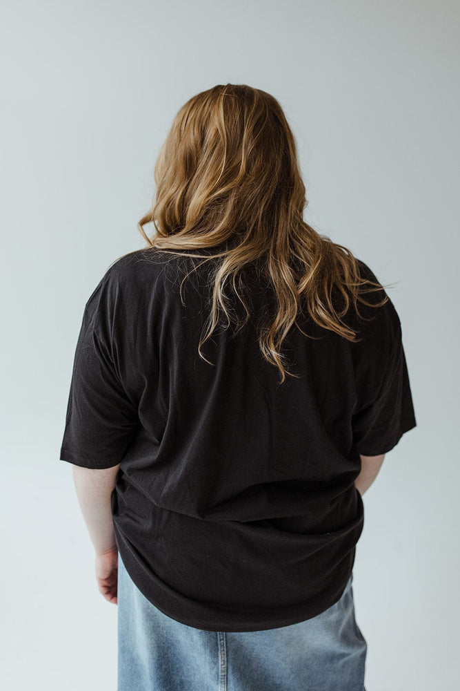 A person with long, wavy hair, wearing the "FARM FRESH PUMPKINS" graphic tee by Love Marlow in black and jeans crafted from super-soft fabric, is standing with their back to the camera, ready for an autumn adventure.