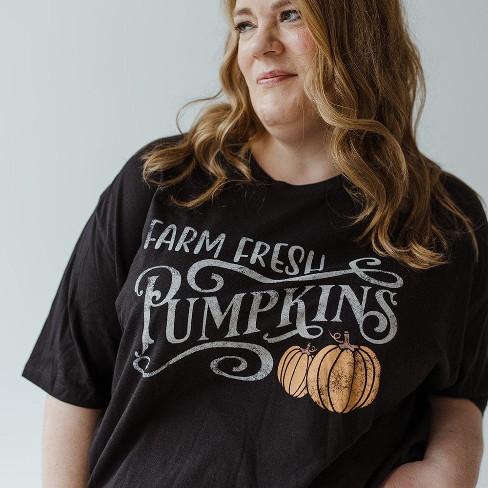 A person with long hair wearing Love Marlow's super-soft "FARM FRESH PUMPKINS" Graphic Tee in Black and a denim skirt gazes to the side against a plain background, embodying the spirit of an autumn adventure.