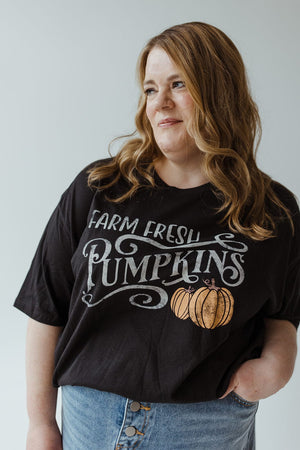 A person with long hair wearing Love Marlow's super-soft "FARM FRESH PUMPKINS" Graphic Tee in Black and a denim skirt gazes to the side against a plain background, embodying the spirit of an autumn adventure.