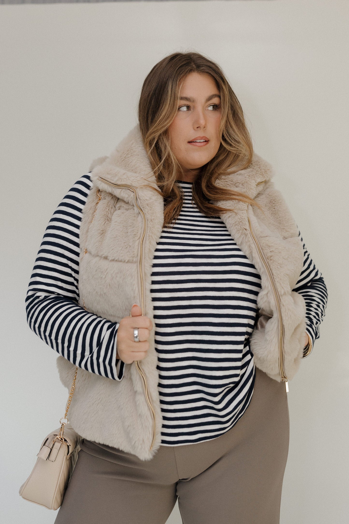 Woman wearing a striped shirt, beige puffer vest, and brown pants against a white background