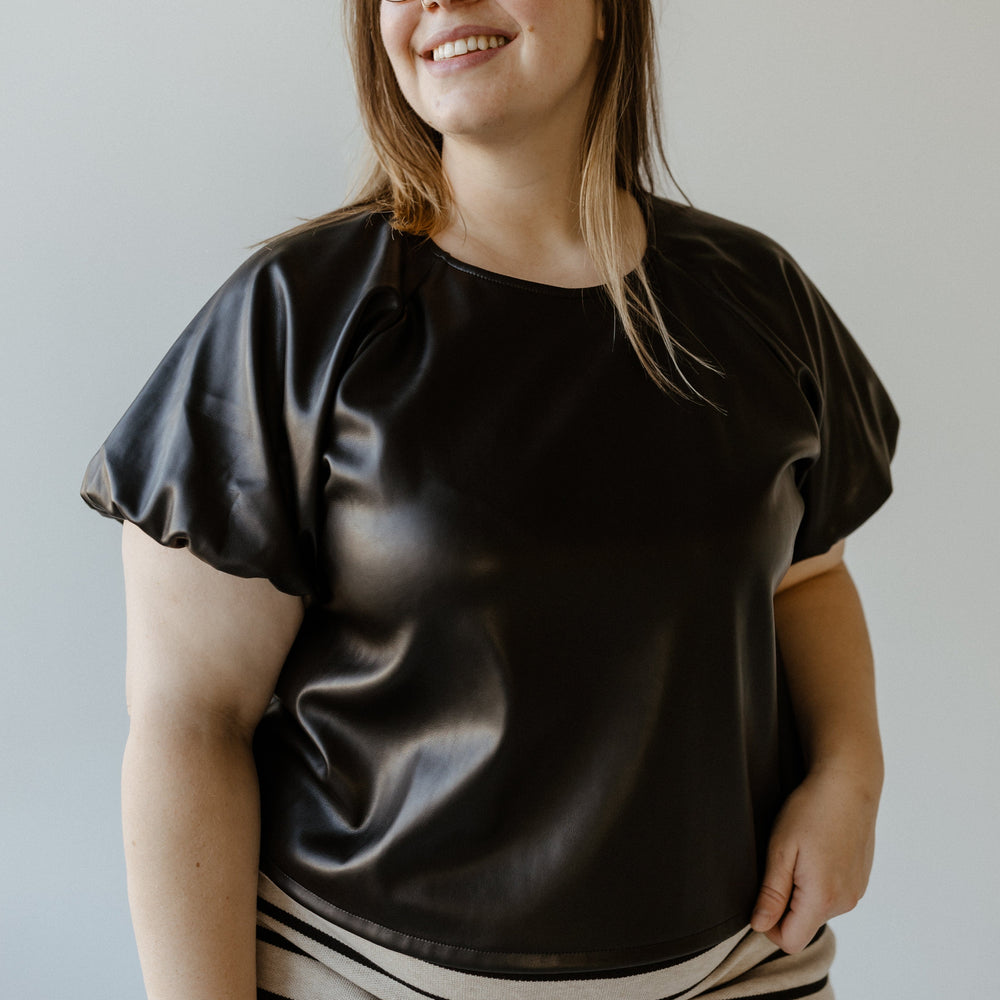 Person wearing a black faux leather blouse and striped skirt, smiling in front of a neutral background.