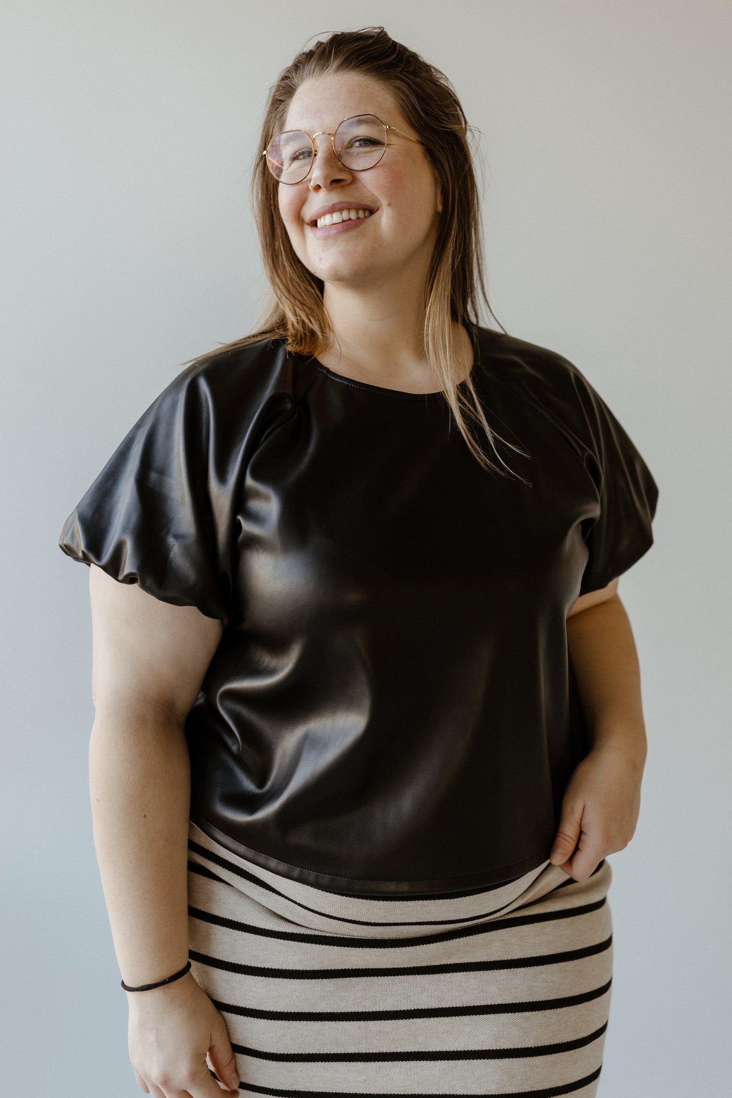 Person wearing a black faux leather blouse and striped skirt, smiling in front of a neutral background.